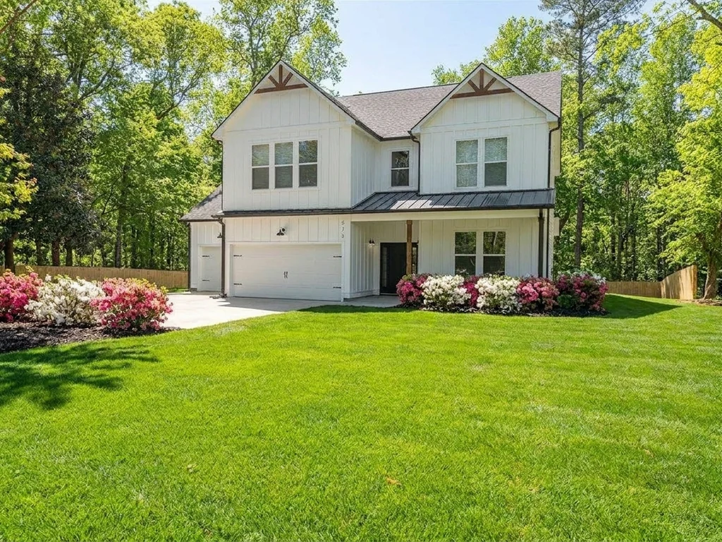 A completed two-story HOC Build home in spring: white board-and-batten siding, dark standing-seam porch roof and timber gable brackets, framed by a mature hardwood with beds of pink and white azaleas along the front.