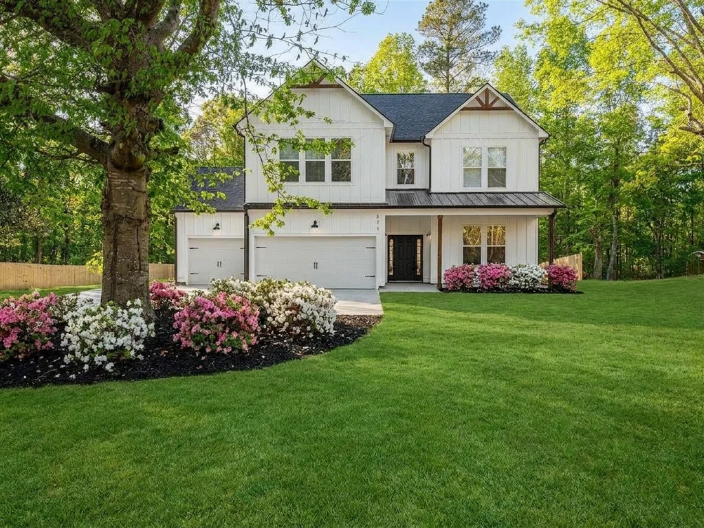Front elevation of the same completed HOC Build home: board-and-batten siding, covered porch with timber posts, three garage bays, azalea beds and a mown lawn.